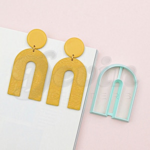 Op de afbeelding: Een paar gele boogvormige oorbellen met een bloemenpatroon en een kleine gele cirkel aan de bovenkant. Een lichtblauwe plastic boogvormige koekjesuitsteker is ook te zien.