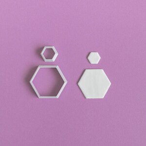 May include: Two white plastic hexagon-shaped cookie cutters on a purple background. The larger cutter is hollow, while the smaller cutter is solid.