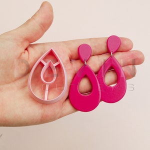 Könnte beinhalten: Ein Paar rosa tropfenförmige Ohrringe mit einem Ausschnittmuster. Die Ohrringe werden neben einer rosa Plastikform zur Herstellung der Ohrringe gezeigt.