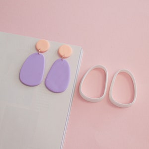 Può includere: Un paio di orecchini color lavanda e pesca a forma di goccia, e un paio di stampi per orecchini ovali bianchi, il tutto su sfondo rosa. Gli orecchini hanno una parte superiore circolare color pesca e una parte inferiore a forma di goccia color lavanda.