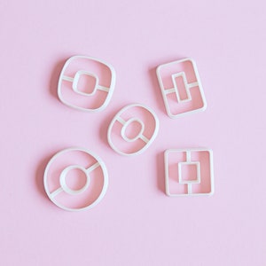 May include: Five white plastic cookie cutters on a pink background. The cutters are shaped like a doughnut, a circle with a line through it, a square with a smaller square inside, and two rectangles with a smaller rectangle inside.