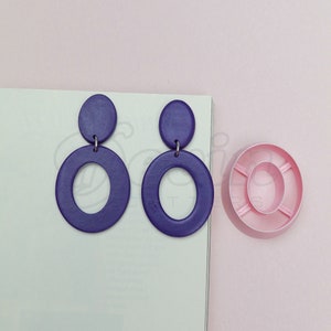 Puede incluir: Un par de pendientes colgantes de color morado con una forma ovalada grande y un círculo más pequeño en la parte superior. Los pendientes están sobre una superficie blanca junto a un objeto circular de color rosa.