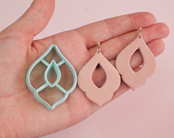 May include: A pair of light pink dangle earrings with a cutout design. The earrings are shown next to a light blue plastic mold of the earring design.