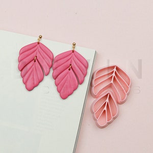 Puede incluir: Pendientes rosas en forma de hoja con detalles dorados. Se muestran junto a un cortador en forma de hoja, sobre un fondo rosa claro. Los pendientes tienen un diseño en capas con vetas detalladas.