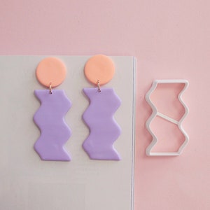 Puede incluir: Un par de pendientes de arcilla polimérica de color morado y melocotón con un diseño ondulado. Los pendientes se muestran junto a un cortador de galletas de plástico blanco con el mismo diseño ondulado.