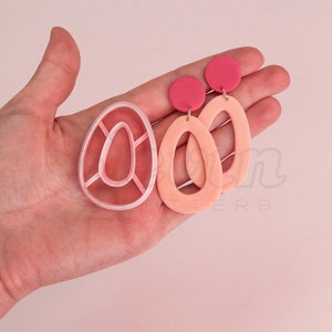 Könnte beinhalten: Ein Paar ovale Ohrringe in Rosa und Pfirsich mit einem kleinen rosa Kreis oben. Die Ohrringe werden neben einer Plastikform zur Herstellung der Ohrringe gezeigt.