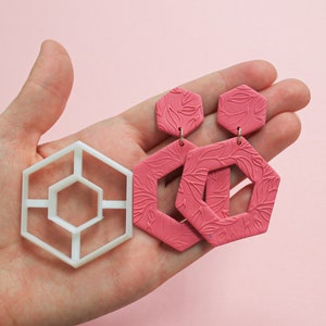 Può includere: Un paio di orecchini esagonali rosa con un motivo a foglia e uno stampo esagonale in plastica bianca.