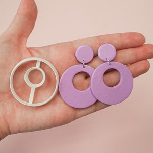 以下が含まれることがあります： 中心に小さな円がくり抜かれた大きな円のデザインのラベンダー色のイヤリングのペア。イヤリングは、イヤリングを作るための白いプラスチック製の型と一緒に示されています。