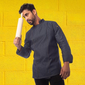 May include: A person wearing a dark blue denim chef jacket with a white rolling pin in their hand. The person is standing in front of a yellow brick wall.