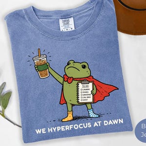 May include: Blue jean t-shirt featuring a cartoon frog superhero holding a coffee and a to-do list. The frog wears a red cape, bow tie, and mismatched socks. Text at the bottom reads "WE HYPERFOCUS AT DAWN."