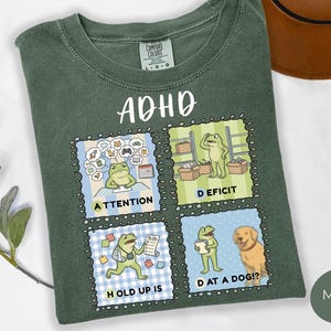 Peut inclure: T-shirt vert mousse avec le mot "ADHD" en écriture blanche. Le t-shirt présente quatre carrés illustrés avec un personnage de grenouille, chacun représentant un aspect différent du TDAH. Les carrés incluent les mots "Attention", "Deficit", "Hold Up Is" et "Dat at a Dog?"
