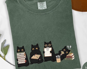 Op de afbeelding: Een mosgroen t-shirt met een afbeelding van vier zwarte katten die ergotherapeutische taken uitvoeren. De katten zijn afgebeeld met boeken met therapietermen, een pegboard, een klembord met Patient Goals en diverse gebruiksvoorwerpen.