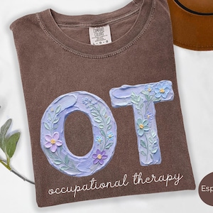May include: Espresso-coloured t-shirt with the letters "OT" in a light blue, floral design. The words "occupational therapy" are written in cursive below the letters. A brown hat is visible in the background.