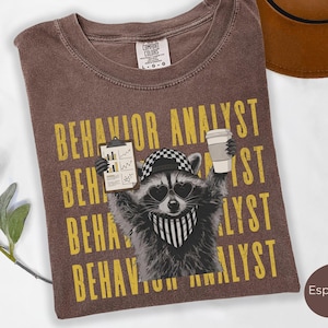 May include: An espresso-coloured t-shirt featuring a graphic of a raccoon wearing heart-shaped sunglasses, a chequered cap, and a striped bandana. The raccoon holds a clipboard and a coffee cup. The text BEHAVIOR ANALYST is repeated in yellow.