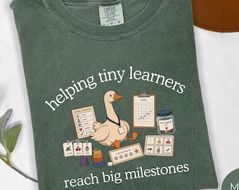 Könnte beinhalten: Ein moosgrünes T-Shirt mit einer Grafik einer Gans, die ein Schlüsselband trägt, umgeben von Lehrmitteln wie einem visuellen Zeitplan, einem Datendiagramm, einem Belohnungsglas und einer Token-Tafel, mit dem Text "helping tiny learners reach big milestones."