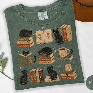 Puede incluir: Una camiseta verde musgo con un estampado gráfico de gatos negros, pilas de libros, una taza y una vela. El diseño incluye libros abiertos con ilustraciones florales y pequeñas estrellas. La camiseta tiene una etiqueta Comfort Colors en el cuello.
