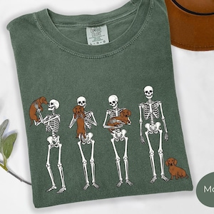 Può includere: Una t-shirt verde muschio con una grafica di tre scheletri bianchi che interagiscono con cani bassotti marroni. Uno scheletro solleva un cane, un altro lo tiene contro il petto, un terzo lo tiene tra le braccia e un cane è seduto a terra.