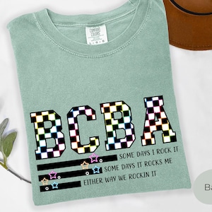 May include: A sage green t-shirt with a crew neckline and a Comfort Colors label. The front features the letters BCBA in a black and white checkered pattern, followed by the text: SOME DAYS I ROCK IT, SOME DAYS IT ROCKS ME, EITHER WAY WE ROCKIN IT.