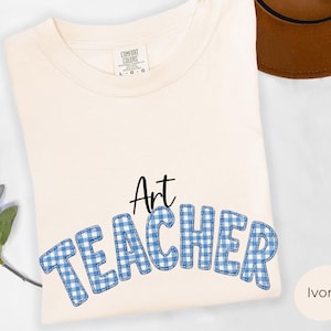 May include: An ivory-colored short-sleeve t-shirt featuring the word Art in black script and the word TEACHER in large, blue and white gingham-patterned block letters with a stitched outline. The shirt has a crew neckline and a Comfort Colors label.