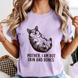 May include: A light purple t-shirt featuring a black and white illustration of a corgi lying on its back. The text "MOTHER, I AM BUT SKIN AND BONES" is printed below the corgi image. The shirt is a solid color.