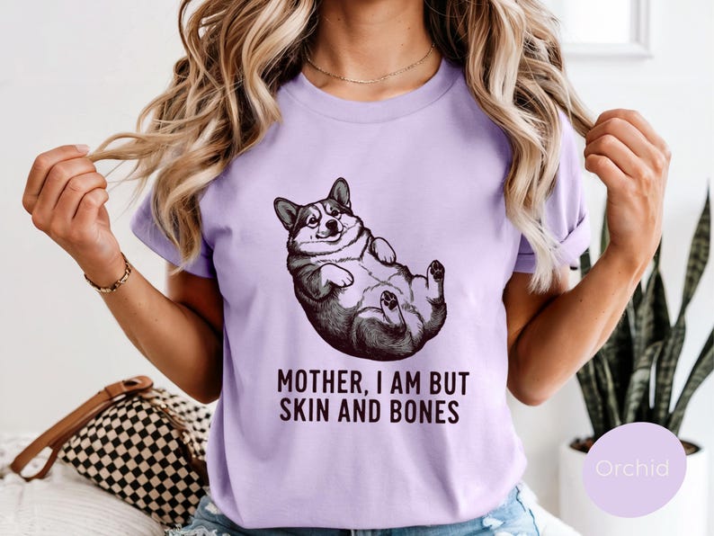 May include: A light purple t-shirt featuring a black and white illustration of a corgi lying on its back. The text "MOTHER, I AM BUT SKIN AND BONES" is printed below the corgi image. The shirt is a solid color.
