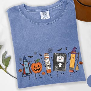 May include: A blue jean colored t-shirt featuring a graphic print of a crayon dressed as a vampire, a jack-o'-lantern, a mummy, a notebook with a ghost, and a pencil wearing a witch hat with a broom. The shirt has a Comfort Colors brand label.