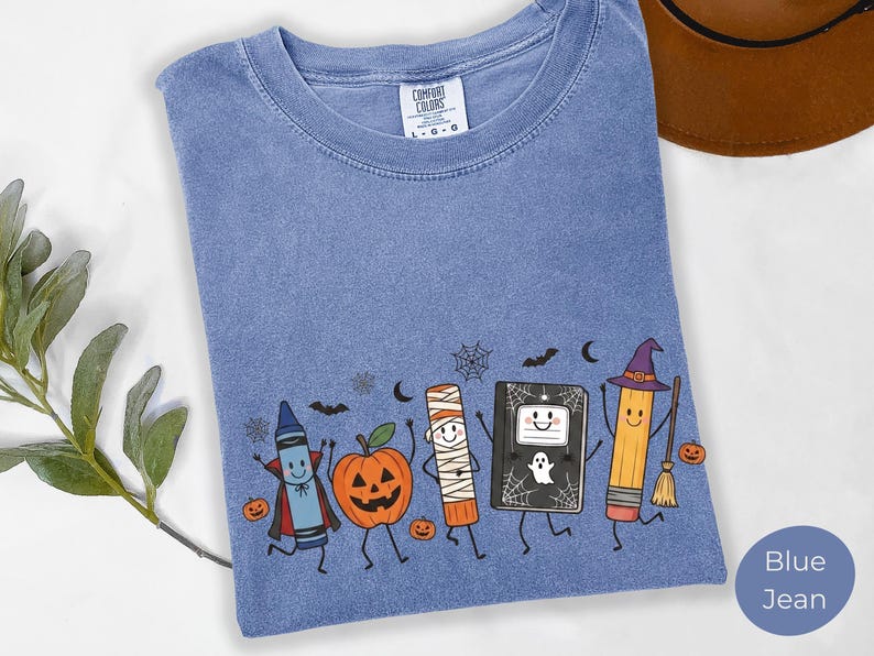 May include: A blue jean colored t-shirt featuring a graphic print of a crayon dressed as a vampire, a jack-o'-lantern, a mummy, a notebook with a ghost, and a pencil wearing a witch hat with a broom. The shirt has a Comfort Colors brand label.