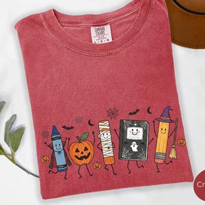 May include: A crimson t-shirt with a graphic print featuring a blue crayon in a cape, a jack-o'-lantern, a mummy, a black notebook with a ghost, and a yellow pencil wearing a witch hat. The shirt has a Comfort Colors label at the neck.