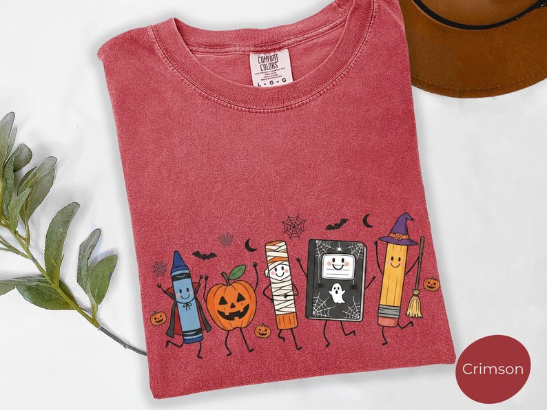 May include: A crimson t-shirt with a graphic print featuring a blue crayon in a cape, a jack-o'-lantern, a mummy, a black notebook with a ghost, and a yellow pencil wearing a witch hat. The shirt has a Comfort Colors label at the neck.