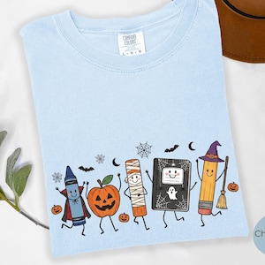 May include: A light blue t-shirt featuring a graphic print of a crayon dressed as a vampire, a jack-o'-lantern, a mummy, a notebook with a ghost, and a pencil dressed as a witch with a broomstick, surrounded by small bats, spiders, and webs.
