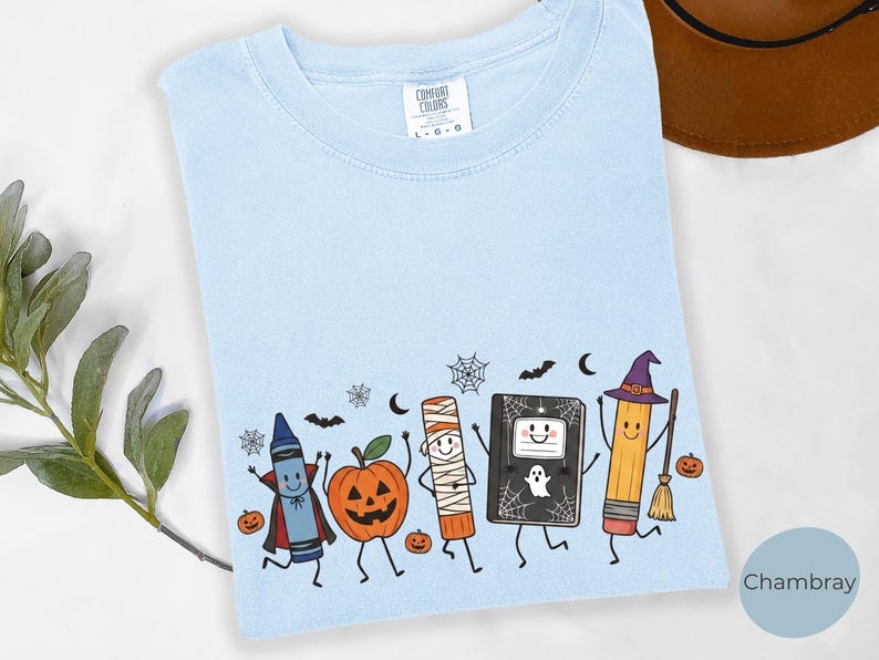 May include: A light blue t-shirt featuring a graphic print of a crayon dressed as a vampire, a jack-o'-lantern, a mummy, a notebook with a ghost, and a pencil dressed as a witch with a broomstick, surrounded by small bats, spiders, and webs.