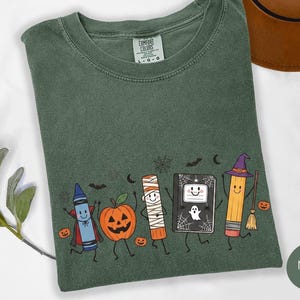 May include: A moss green short-sleeve t-shirt featuring a graphic print of a blue crayon vampire, a jack-o'-lantern, a mummy glue stick, a ghost-adorned notebook, and a pencil wearing a witch hat, all surrounded by small bats and spiderwebs.