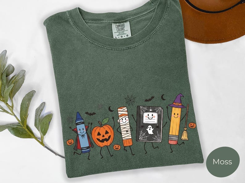 May include: A moss green short-sleeve t-shirt featuring a graphic print of a blue crayon vampire, a jack-o'-lantern, a mummy glue stick, a ghost-adorned notebook, and a pencil wearing a witch hat, all surrounded by small bats and spiderwebs.