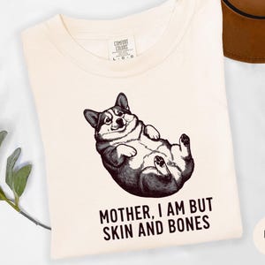 May include: Ivory-colored t-shirt featuring a black and white illustration of a corgi lying on its back. The text "MOTHER, I AM BUT SKIN AND BONES" is printed below the corgi. A brown hat is visible in the background.