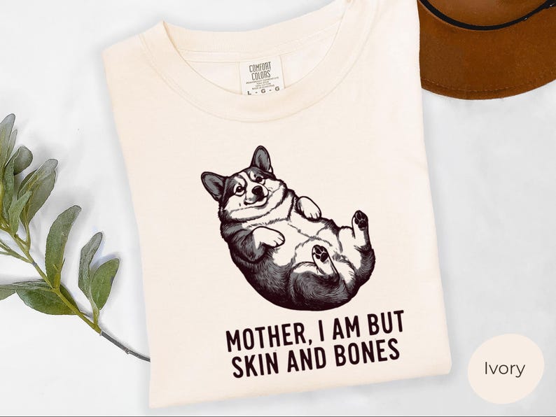 May include: Ivory-colored t-shirt featuring a black and white illustration of a corgi lying on its back. The text "MOTHER, I AM BUT SKIN AND BONES" is printed below the corgi. A brown hat is visible in the background.