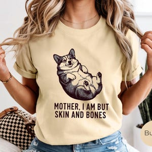 May include: A light yellow t-shirt featuring a black and white illustration of a corgi lying on its back. The text "MOTHER, I AM BUT SKIN AND BONES" is printed below the corgi image. The shirt has short sleeves.