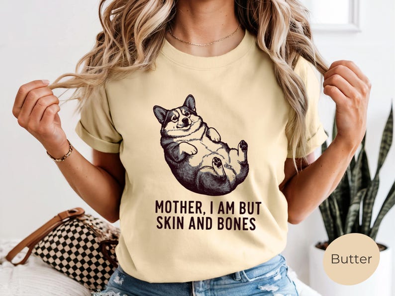 May include: A light yellow t-shirt featuring a black and white illustration of a corgi lying on its back. The text "MOTHER, I AM BUT SKIN AND BONES" is printed below the corgi image. The shirt has short sleeves.
