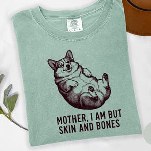May include: A light green t-shirt featuring a graphic of a corgi lying on its back. The corgi is in a relaxed pose with its paws in the air. The text "MOTHER, I AM BUT SKIN AND BONES" is printed below the corgi in a bold font.