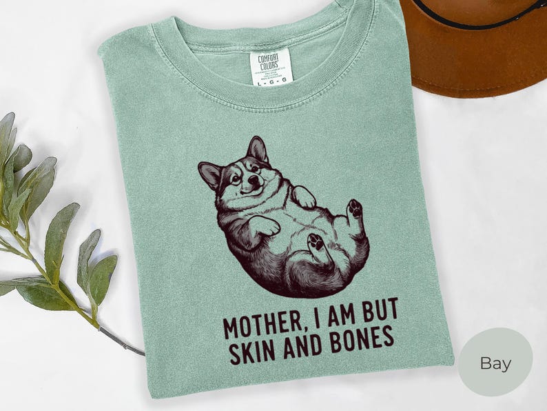 May include: A light green t-shirt featuring a graphic of a corgi lying on its back. The corgi is in a relaxed pose with its paws in the air. The text "MOTHER, I AM BUT SKIN AND BONES" is printed below the corgi in a bold font.