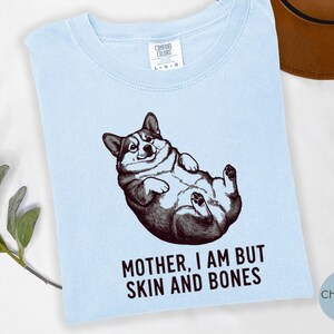 May include: Light blue t-shirt featuring a detailed illustration of a corgi lying on its back. The graphic is in black and white, with the text "MOTHER, I AM BUT SKIN AND BONES" below the image. The shirt has a crew neck.