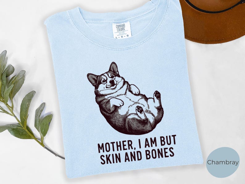 May include: Light blue t-shirt featuring a detailed illustration of a corgi lying on its back. The graphic is in black and white, with the text "MOTHER, I AM BUT SKIN AND BONES" below the image. The shirt has a crew neck.