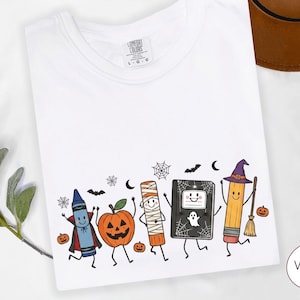 May include: A white t-shirt featuring a colorful graphic of cartoon characters dressed for Halloween, including a crayon in a vampire cape, a jack-o'-lantern, a mummy, a notebook with a ghost, and a pencil with a witch hat and broomstick.