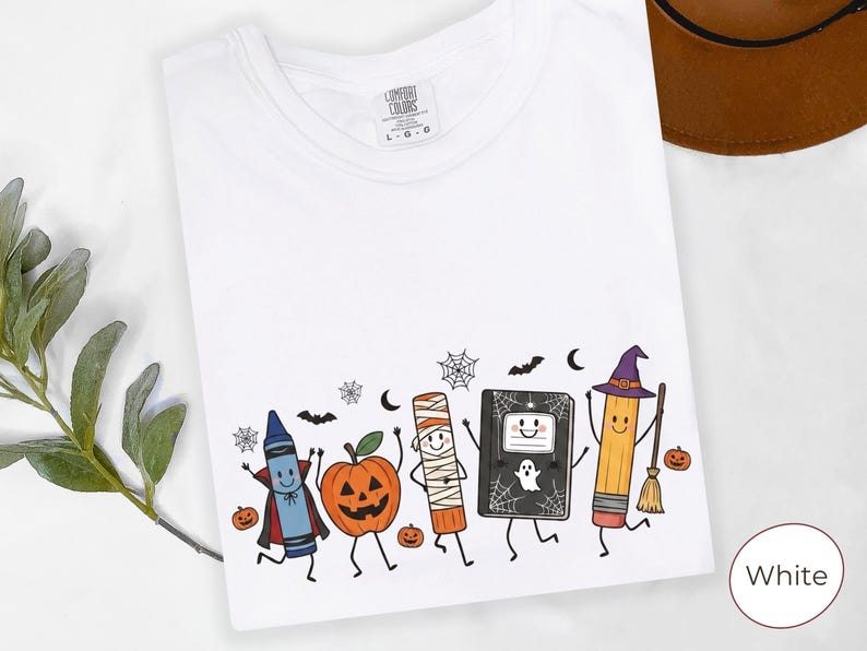 May include: A white t-shirt featuring a colorful graphic of cartoon characters dressed for Halloween, including a crayon in a vampire cape, a jack-o'-lantern, a mummy, a notebook with a ghost, and a pencil with a witch hat and broomstick.