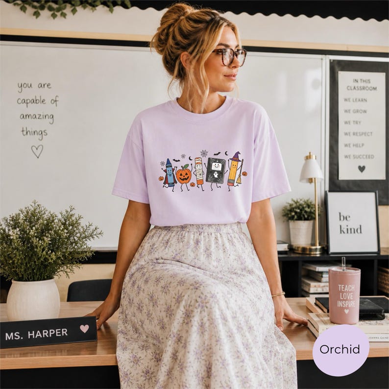 May include: A light purple short-sleeve t-shirt featuring a graphic print of a smiling crayon, a jack-o'-lantern, a glue stick, a ghost in a book, and a pencil wearing a witch hat, all surrounded by small bats and snowflakes.