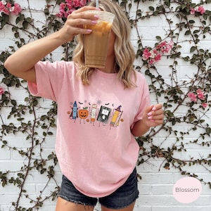 May include: A light pink short-sleeve t-shirt featuring a graphic print of a blue crayon, an orange jack-o'-lantern, a mummy, a black book with a ghost, and a yellow pencil with a witch hat, all surrounded by small bats and stars.