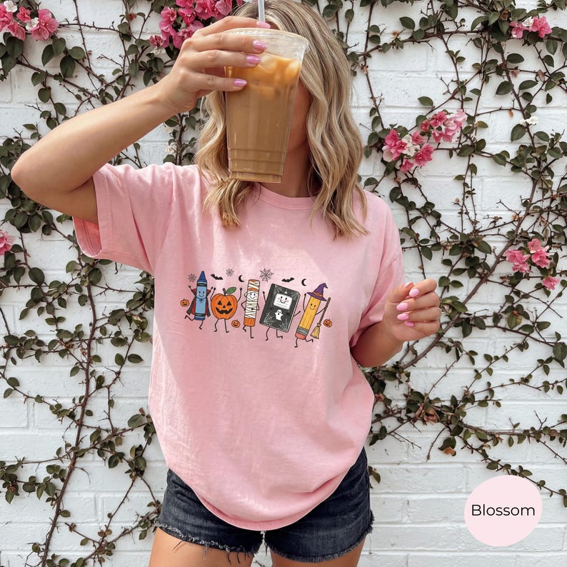 May include: A light pink short-sleeve t-shirt featuring a graphic print of a blue crayon, an orange jack-o'-lantern, a mummy, a black book with a ghost, and a yellow pencil with a witch hat, all surrounded by small bats and stars.