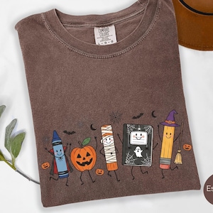 May include: An espresso-colored t-shirt featuring a graphic print of a blue crayon vampire, a jack-o'-lantern, a mummy, a black notebook with a ghost, and a pencil wearing a witch hat. The shirt has a crew neck and a Comfort Colors brand label.