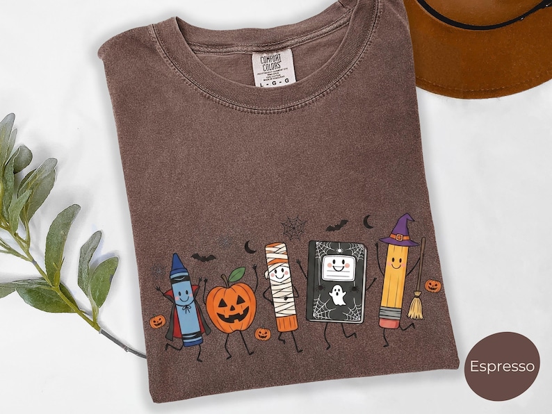 May include: An espresso-colored t-shirt featuring a graphic print of a blue crayon vampire, a jack-o'-lantern, a mummy, a black notebook with a ghost, and a pencil wearing a witch hat. The shirt has a crew neck and a Comfort Colors brand label.