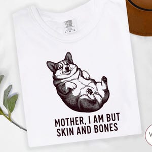 May include: White t-shirt featuring a black and white illustration of a corgi lying on its back. The text "MOTHER, I AM BUT SKIN AND BONES" is printed below the corgi. The shirt has a crew neck and short sleeves.