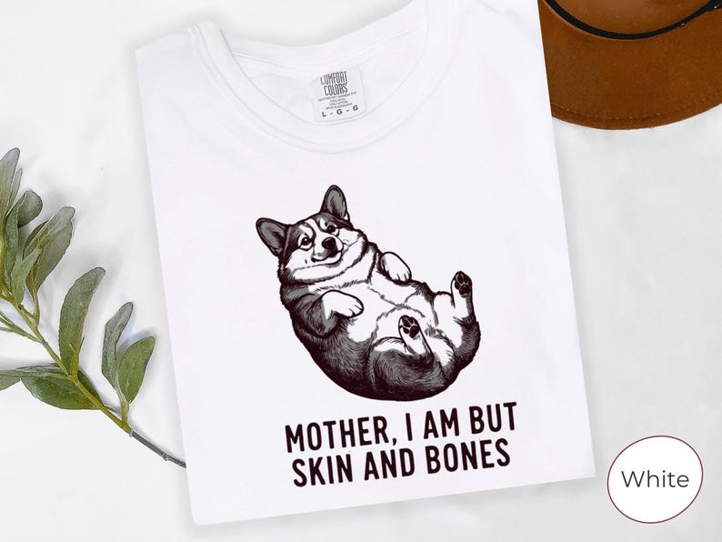 May include: White t-shirt featuring a black and white illustration of a corgi lying on its back. The text "MOTHER, I AM BUT SKIN AND BONES" is printed below the corgi. The shirt has a crew neck and short sleeves.